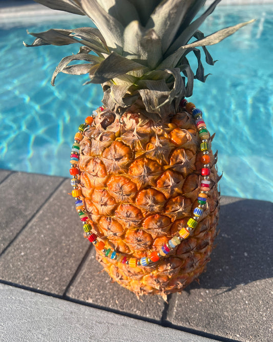 A whole pineapple standing upright on a pool deck, wrapped in colorful beaded necklaces and a beaded waist wrap, with a swimming pool in the background.