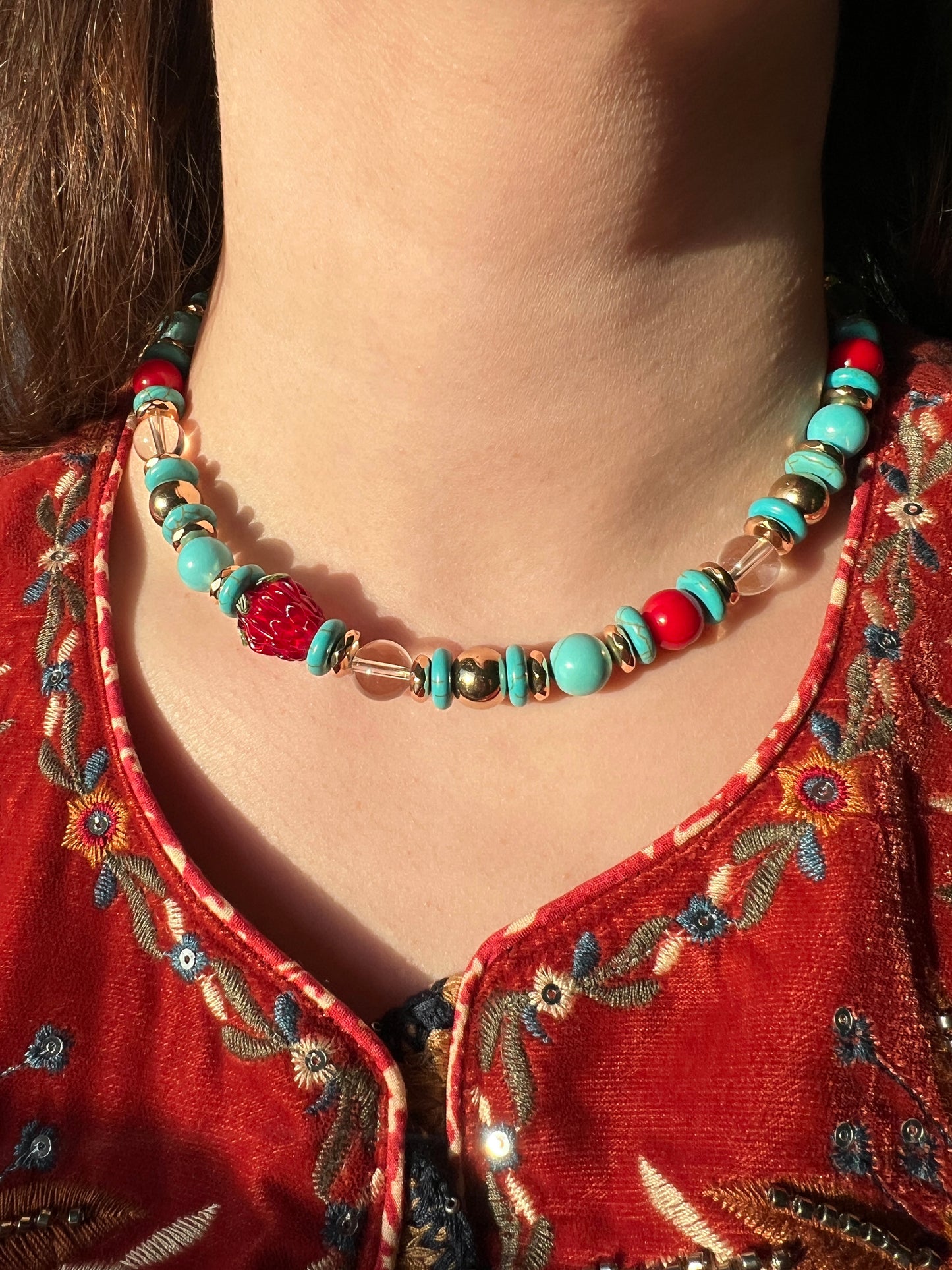 Necklace with turquoise and red beads worn by a person in an embroidered red garment.