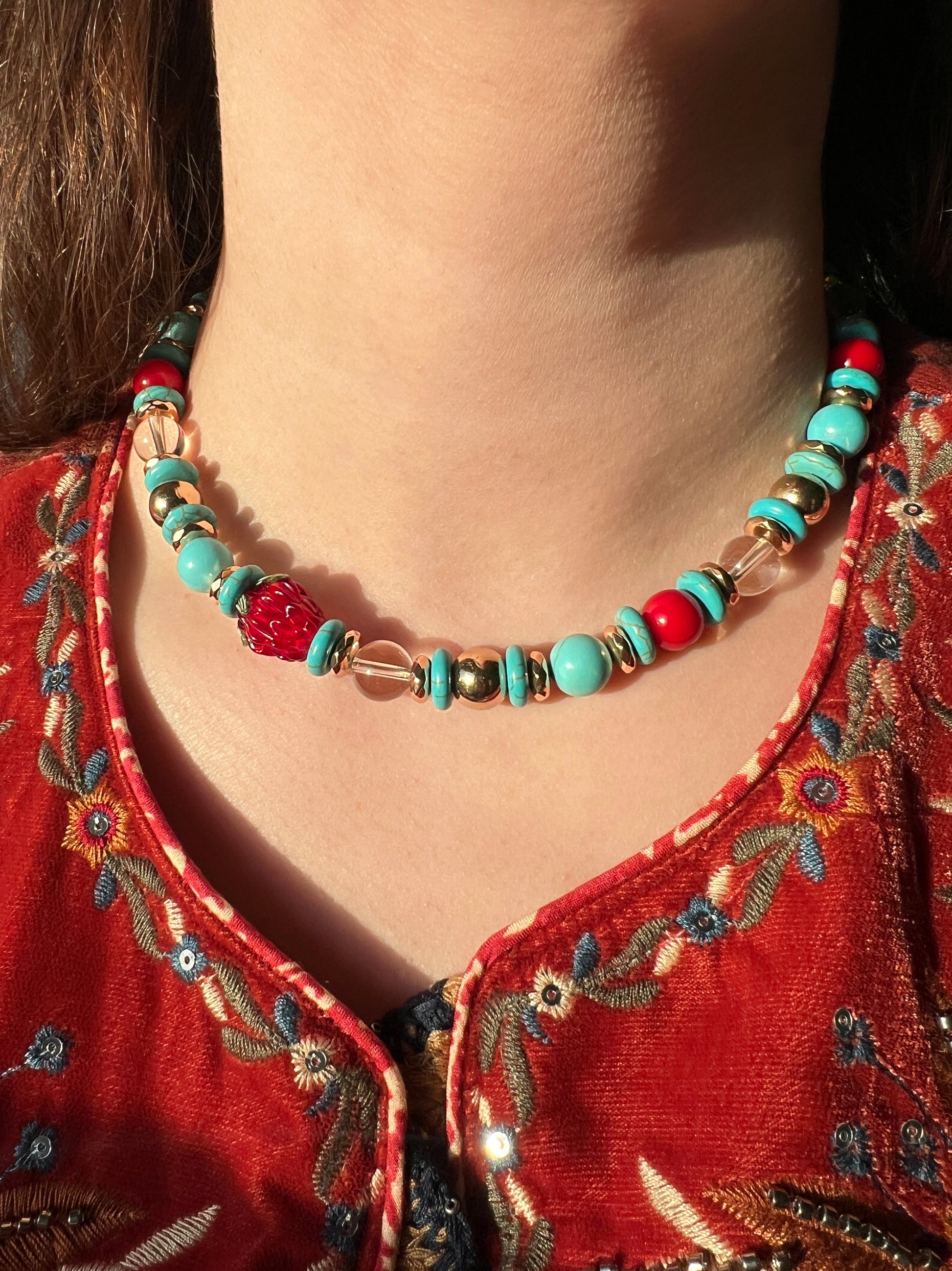 Necklace with turquoise and red beads worn by a person in an embroidered red garment.