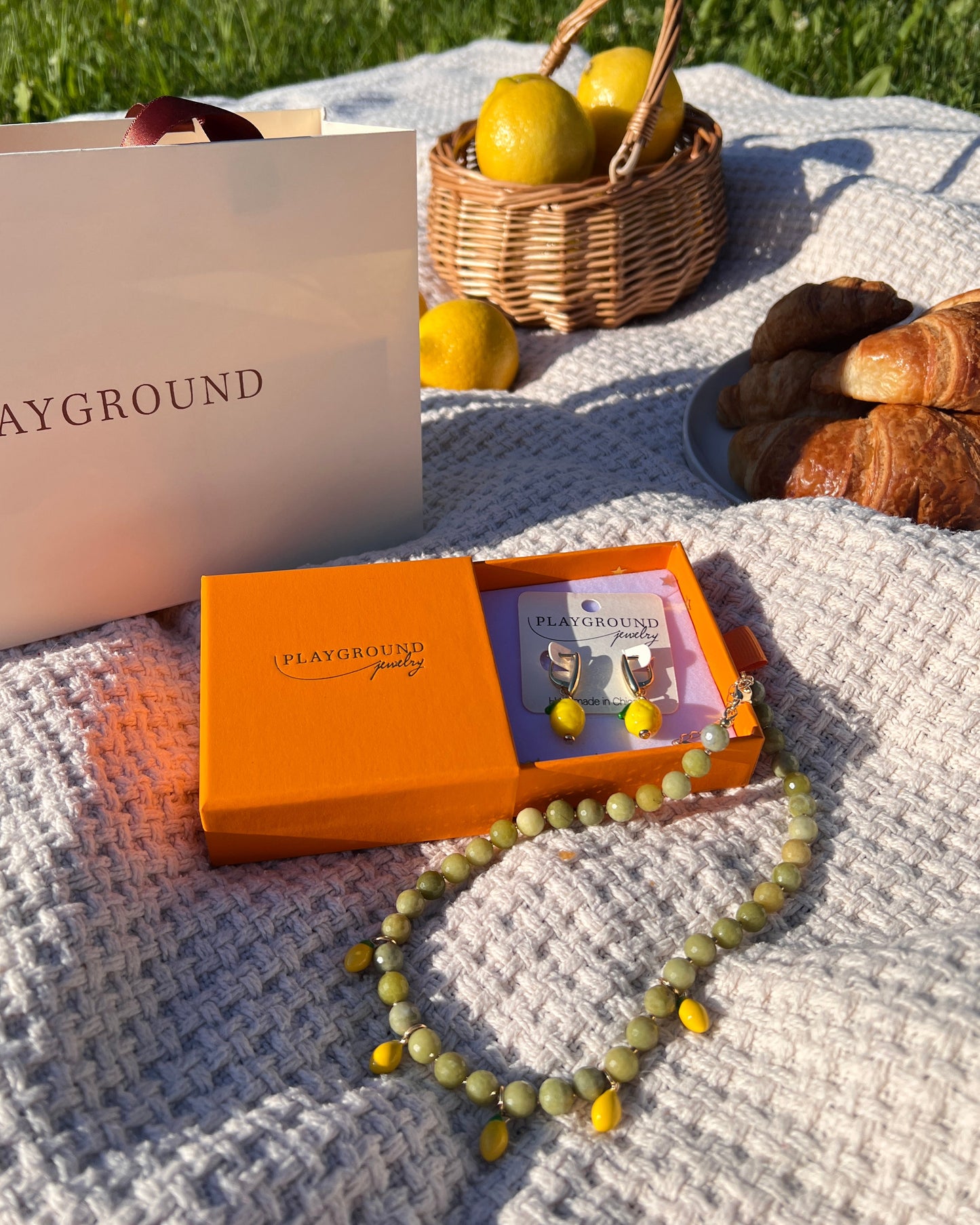 Orange box, bag titled 'PLAYGROUND', basket with lemons, and necklace on a blanket outdoors.