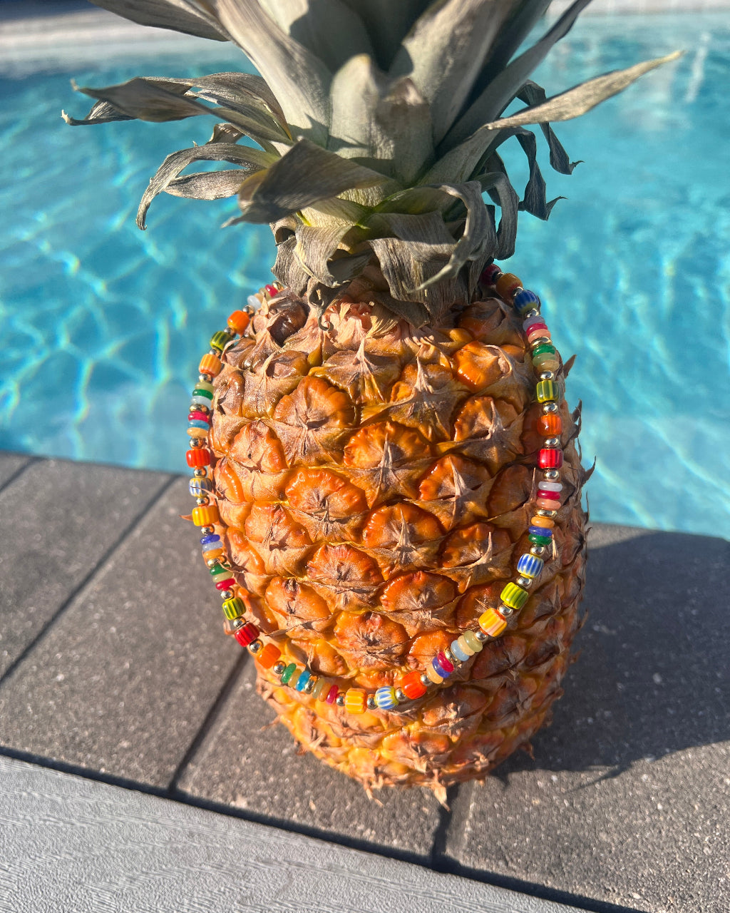 A whole pineapple standing upright on a pool deck, wrapped in colorful beaded necklaces and a beaded waist wrap, with a swimming pool in the background.