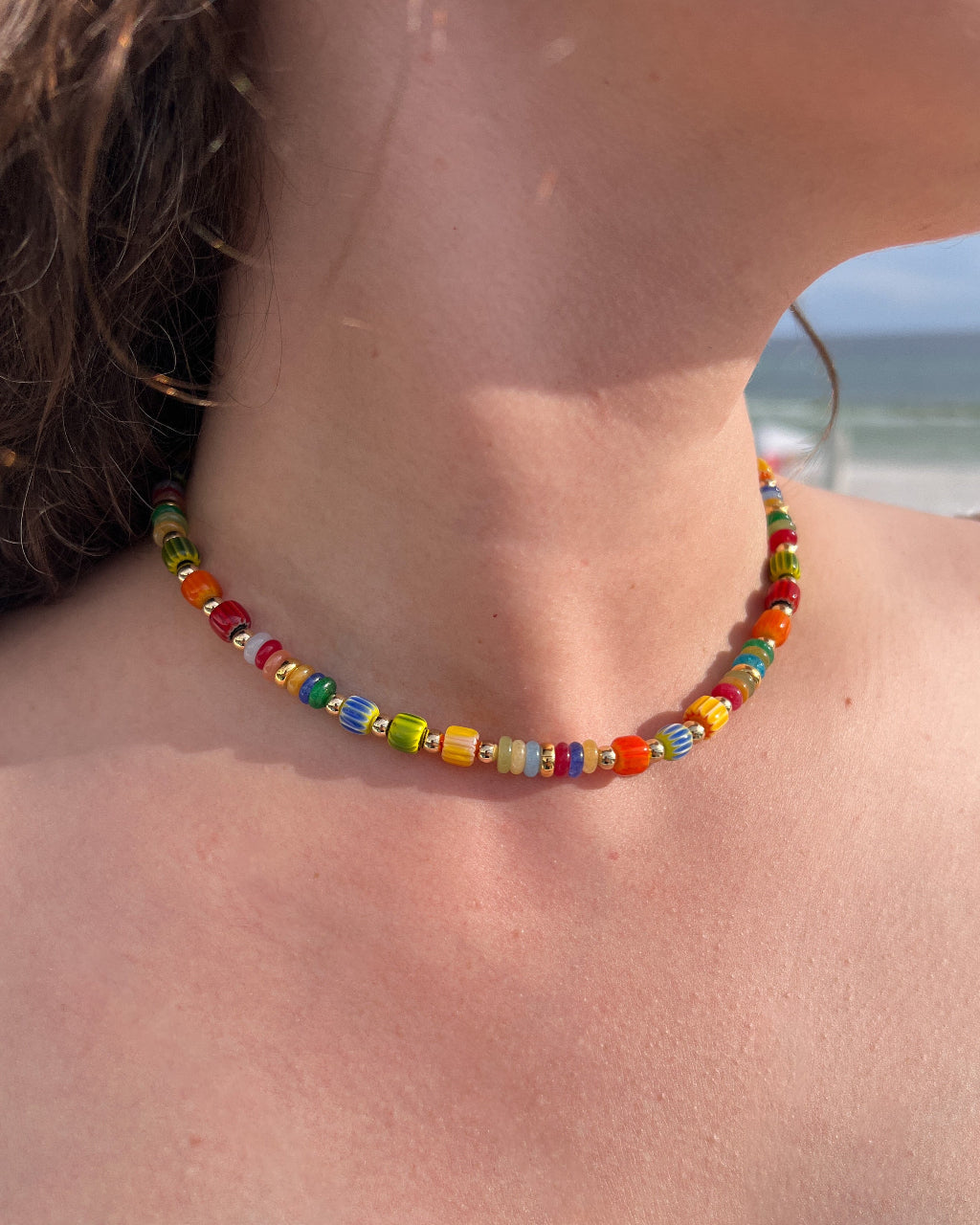 A close-up lifestyle shot of a person wearing a colorful beaded choker necklace at the beach, with ocean waves visible in the background.